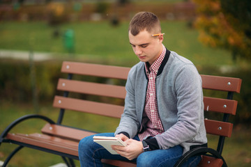 Obraz premium A cute student sits on the bench and writes down his thoughts in his notebool using a pensil. study outside