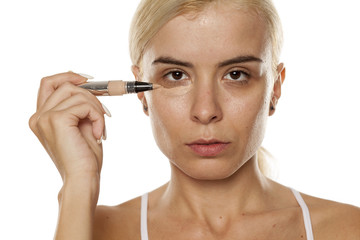 Young woman applying concealer under her eyes with brush on white background
