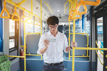 Young Asian man traveler standing on a bus using smartphone watch video or playing game while smile of happy, transport, tourism and road trip concept