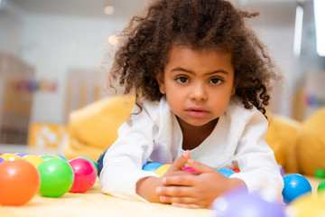 adorable african american kid lying on carpet with toys in kindergarten and looking at camera