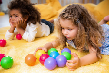 Fototapeta na wymiar adorable multiethnic kids playing on yellow carpet with colored balls in kindergarten
