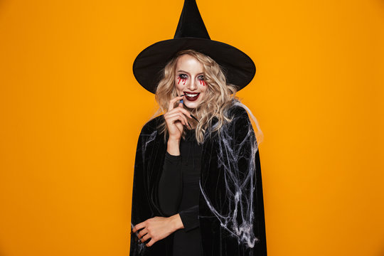 Image of tricky witch woman wearing black costume and halloween makeup looking at camera, isolated over yellow background