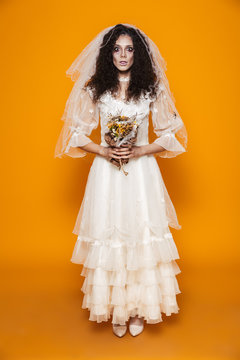 Full length image of dead bride zombie on halloween wearing wedding dress and scary makeup holding flowers, isolated over yellow background