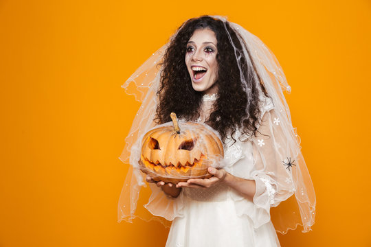 Image of spooky bride zombie on halloween wearing wedding dress and scary makeup holding carved pumpkin, isolated over yellow background
