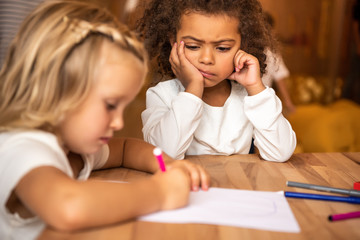 dissatisfied african american kid looking at caucasian child drawing at table in kindergarten