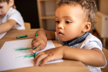 adorable african american boy drawing with green felt tip pen in kindergarten