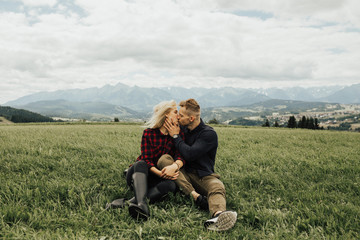 Naklejka na ściany i meble Couple sitting and kissing on grass on the top of mountains enjoying mountains and clouds landscape. Love and travel concept. Lifestyle concept. family active traveling. Mountain view
