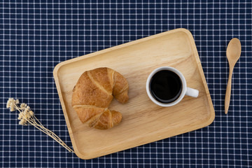 A cup of coffee and croissant with white flower and wooden spoon on dark blue table cloth