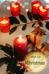 Merry Christmas greeting card with text. Lit candles, gift boxes and branch of holly