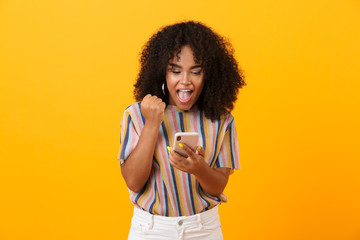 Emotional african woman posing isolated over yellow background using mobile phone make winner...