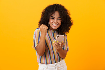 Emotional african woman posing isolated over yellow background using mobile phone make winner...