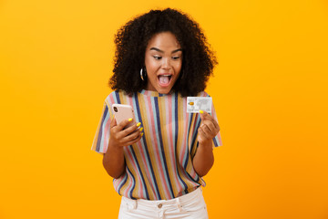 Excited happy african woman posing isolated over yellow background using mobile phone holding credit card.