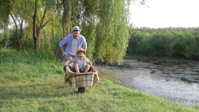 happy family vacation in countryside, father fools around with his son in a wheelbarrow near river with reeds
