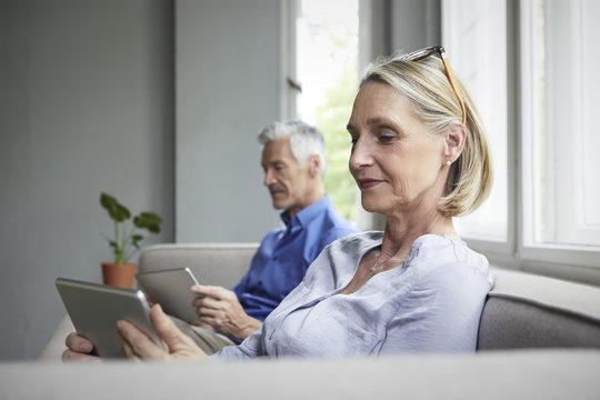 Mature couple sitting on couch at home using tablets