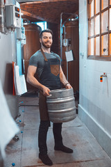 A young brewer in an apron holds a barrel with beer in the hands of a brewery