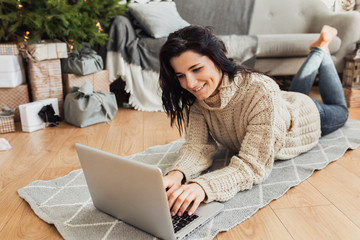 Image of beautiful smiling young woman shopping online on laptop in cozy Christmas interior. Female lying on the floor next the Christmas tree and sofa, bying gifts for the family on winter sales.