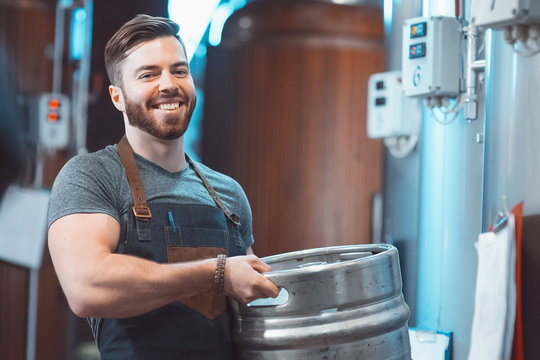 A young brewer in an apron holds a barrel with beer in the hands of a brewery