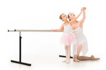 happy female teacher in tutu helping little ballerina exercising at ballet barre stand isolated on white background