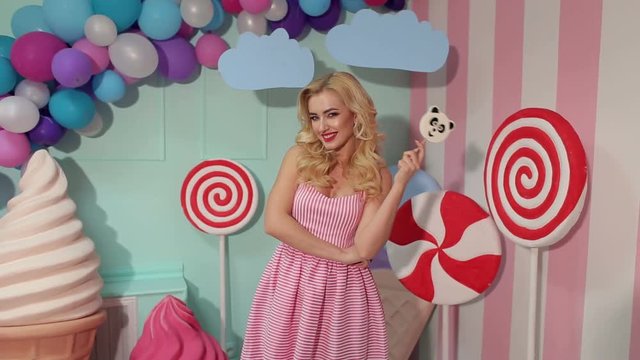 Portrait of a cheerful and happy girl in a pink striped dress with a large candy in the form of a panda on the background of colorful balloons, a large plastic ice cream and candy.
