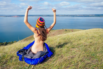 Back view of sexy naked woman tourist in a funny hat from Nepal sitting in blue sleeping bag on the grass, enjoying sunny morning in the mountains lake. - obrazy, fototapety, plakaty