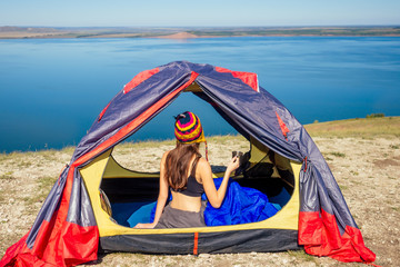 Back view of sexy naked woman tourist in a funny hat from Nepal sitting in blue sleeping bag in a tent holding a cup of tea, coffee sunny good morning in the mountains lake.