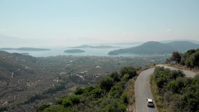 Aerial - Car driving on a road that rises above the city of Nidri
