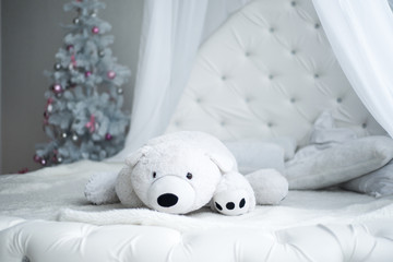 modern child's bedroom decorated for Christmas: a Toy polar bear lying on a white bed, on the background of Christmas tree
