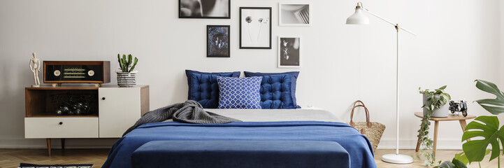Real photo of a blue bed, retro cabinet, art gallery and lamp in a bedroom interior