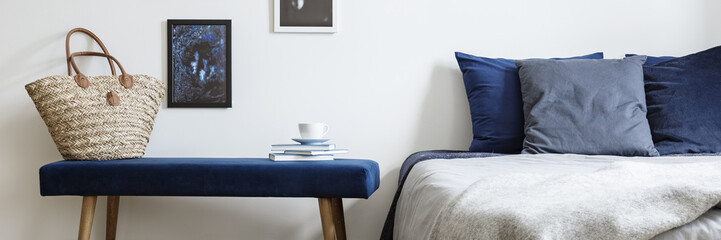 Close-up of a bench with a wicker bag and cup on books next to a bed in a bedroom interior. Real photo