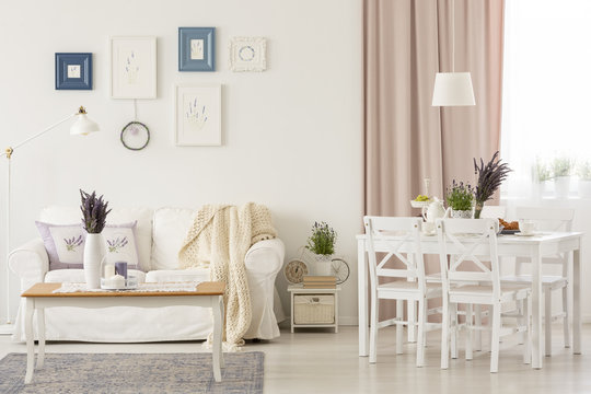 Flowers on table in white living room interior with pink drapes and posters above sofa. Real photo