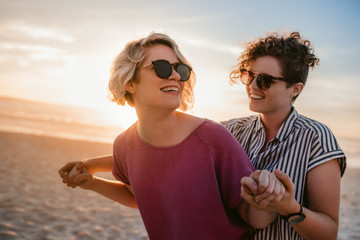 Laughing lesbian couple dancing playfully on a beach at sunset - Powered by Adobe