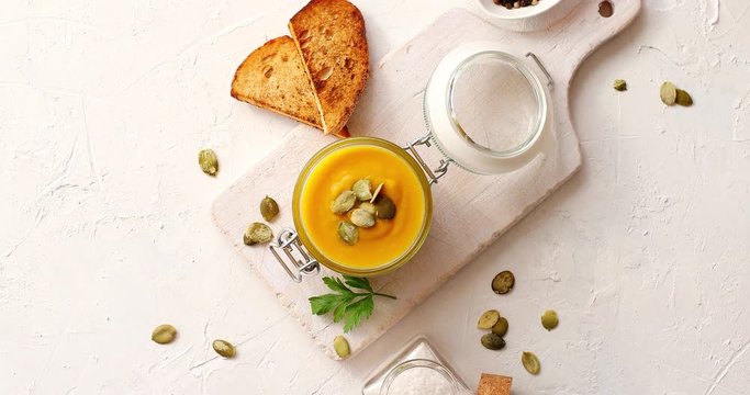 From above view of yellow pumpkin soup served in jar on wooden chopping board with bread and bowl of spices laid near on white background