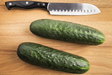 two whole green cucumbers next to a knife on a wooden table
