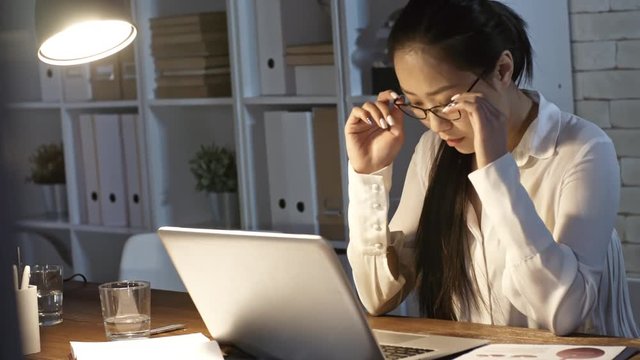 Young asian businesswoman taking off eyeglasses and rubbing tired eyes while working on laptop late in the office