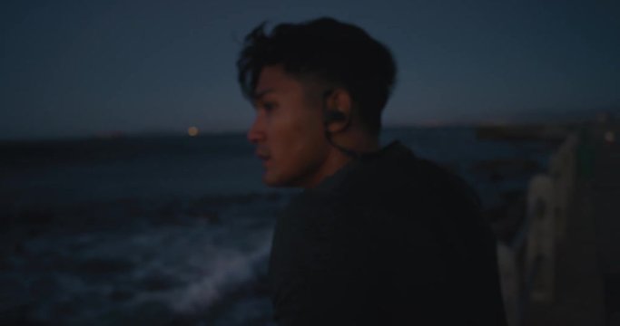 Young Asian Man Runner Resting After Running Workout Exhausted Athlete Takes Off Earphones Looking Serious At Camera On Calm Seaside At Sunset Close Up Rear View