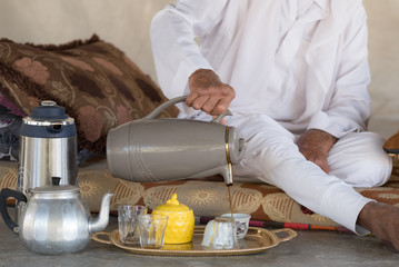 Arab Bedouin Man sits on the floor and pours tea or coffee from teapot into the white cup in Bedouin tent.White traditional Arabic clothing.