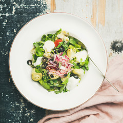 Flat-lay of fresh green summer salad with artichokes, olives, soft cheese and red onion in white plate over rustic wooden painted background, top view, square crop