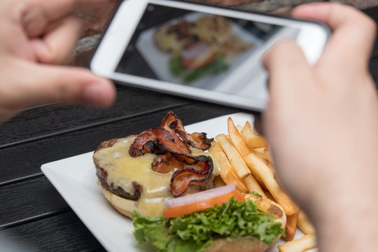 Man takes photo of food with mobile phone at an outdoor bar.  Taking a picture of your food with your phone. Hamburger, fries, and beer on a white plate outside on a black table, POV shot.
