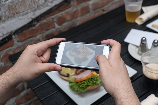 Point of view shot, man takes photo of food with mobile phone at an outdoor bar.  Taking a picture of your food with your phone. Hamburger, fries, and beer on a white plate outside on a black table.