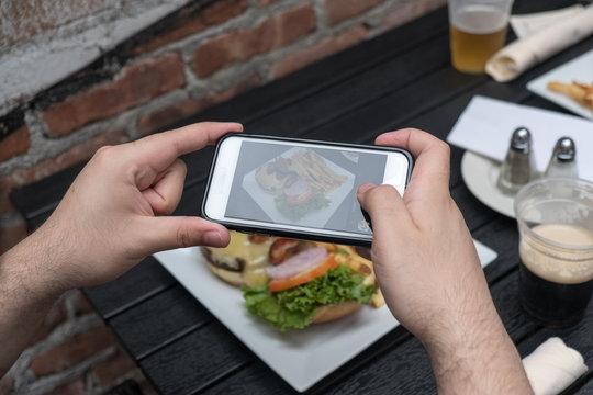 Point of view shot, man takes photo of food with cell phone at an outdoor bar.  Taking a picture of your food with your phone. Hamburger and fries on a white plate outside on a black table.