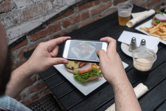 Point of view shot, man takes photo of food with mobile phone at an outdoor bar.  Taking a picture of your food with your phone. Hamburger and fries on a white plate outside on a black table.