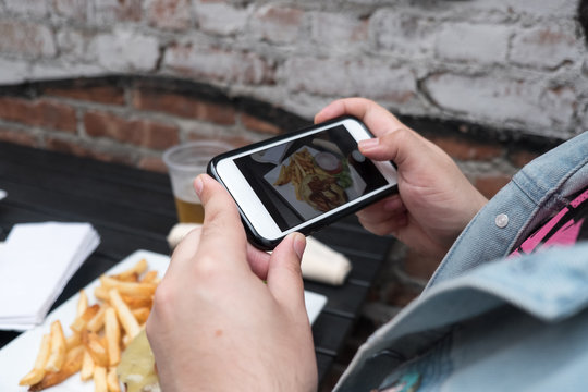 Man holding a mobile phone taking a photo of his food. Cell phone food photography. Taking a picture of hamburger, beer, and french fries at an outdoor pub with a smartphone.