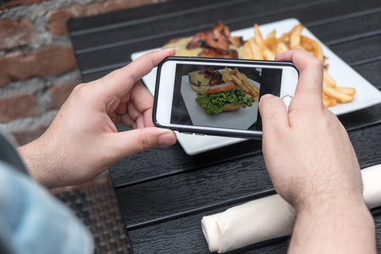 Man takes a photo of his lunch with his cell phone. Mobile phone photography. Taking a photo with phone of hamburger, french french fries on white plate on black table outside.