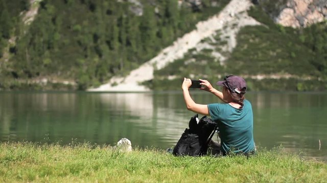 Young woman uses a smartphone to take a picture of beautiful view by the lake, Lago Di Braies in Italy
