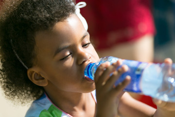 A black child eagerly drinks water from a bottle. A little girl keeps a thirst out of a plastic bottle on a hot day.