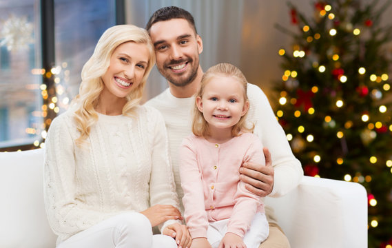 family, holidays and people concept - happy mother, father and little daughter sitting on sofa at home over christmas tree lights background