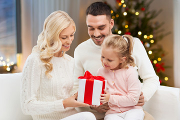 Fototapeta na wymiar family, holidays and people concept - happy mother, father and little daughter with gift box sitting on sofa at home over christmas tree lights background