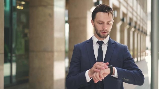 Smiling young businessman walking in a summer street and making a stop to look at his smart watch. Tracking slow motion low medium shot