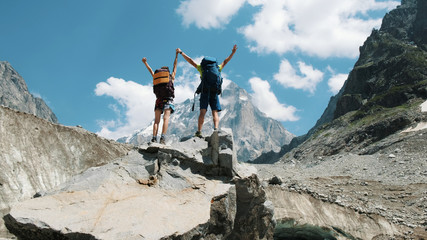 Couple of tourists with backpacks in the campaign raise their hands up on the top of the mountains. Concept of victory, achievement of the goal and success