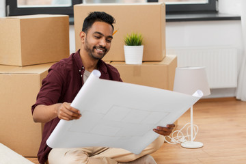 moving, people and real estate concept - happy indian man blueprint and boxes at new home